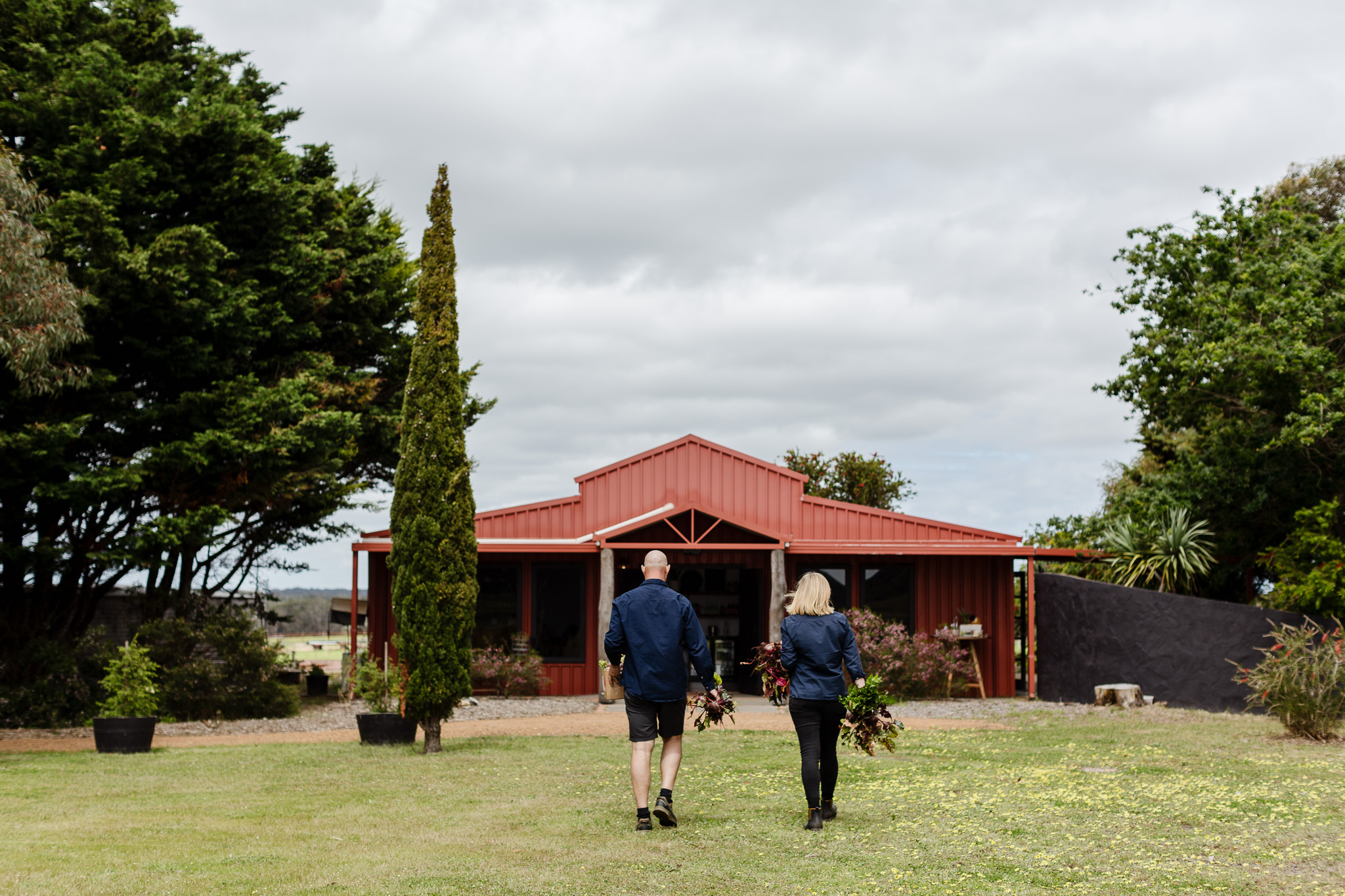 Wild Distillery - Two people walk across a grassy lawn towards a red house, each carrying a bundle of branches or foliage. The sky is cloudy and trees surround the house. - Margaret River WA Wild Distillery Two people walk across a grassy lawn towards a red house, each carrying a bundle of branches or foliage. The sky is cloudy and trees surround the house. Margaret River WA